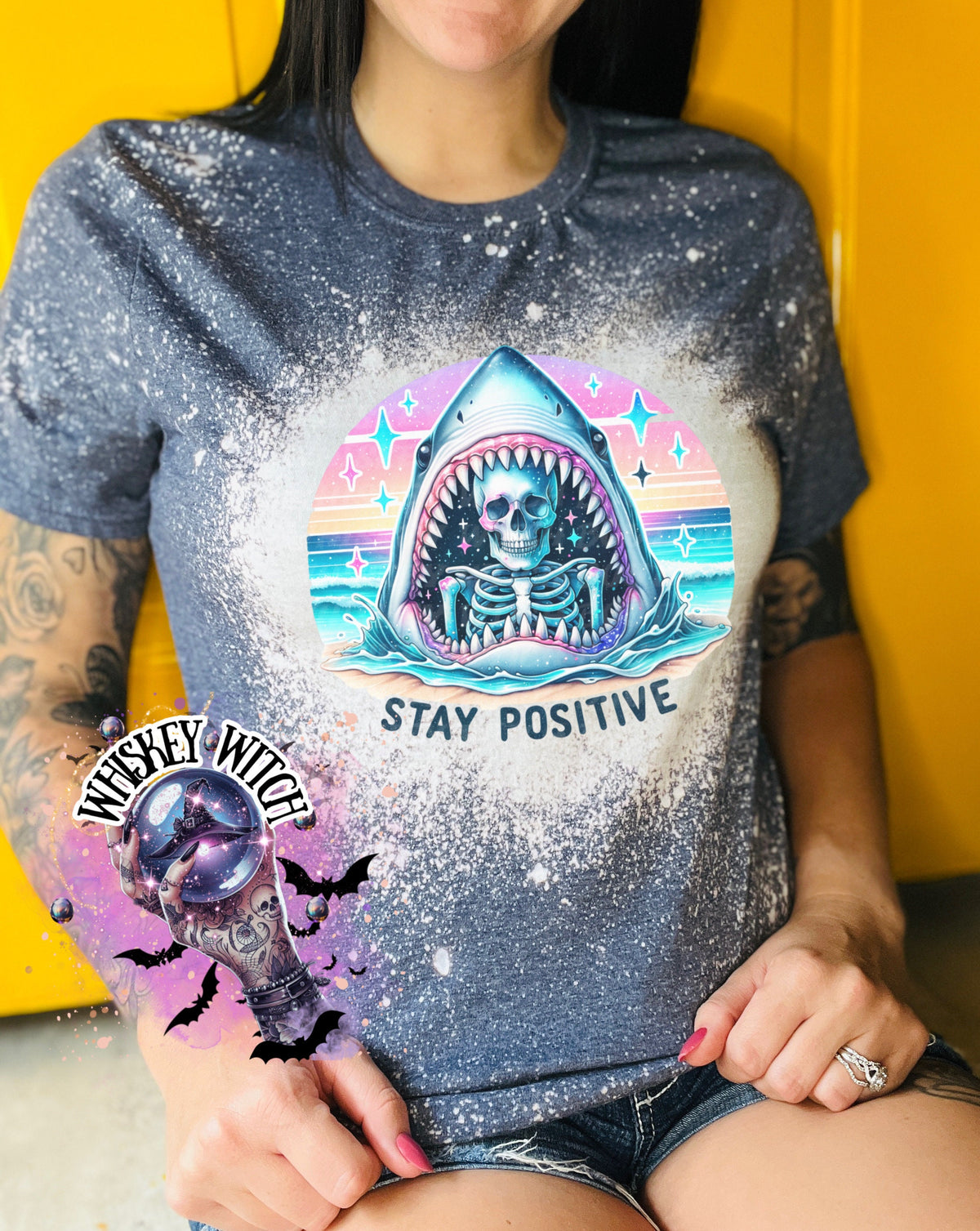 A navy blue  t-shirt with a bleached design of a skeleton inside a shark's mouth. The shark is surrounded by a rainbow and stars. The text "Stay Positive" is below the shark.