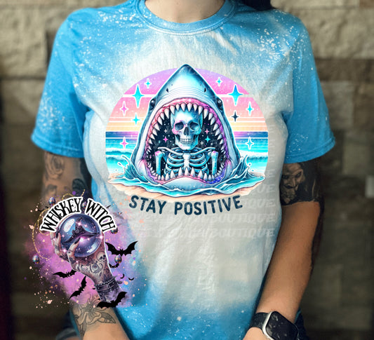 A sapphire blue  t-shirt with a bleached design of a skeleton inside a shark's mouth. The shark is surrounded by a rainbow and stars. The text "Stay Positive" is below the shark.