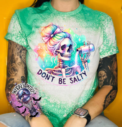 A green t-shirt with a bleached effect featuring a skeleton with rainbow hair pouring salt from a shaker. The text "Don't Be Salty" is printed on the shirt.