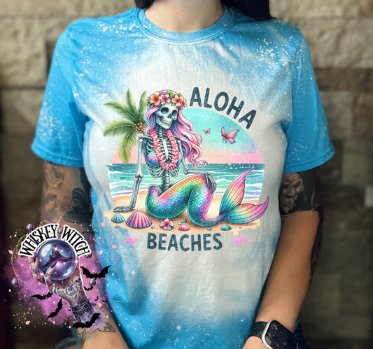 A blue  t-shirt with a bleached effect features a colorful skeleton mermaid with a flower crown, palm tree, and seashells. The text "Aloha Beaches" is printed on the shirt.