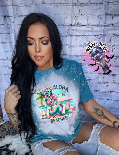 A Galapagos blue bleached t-shirt with a graphic of a skeleton mermaid with pink hair and a lei. The graphic says "Aloha Beaches".