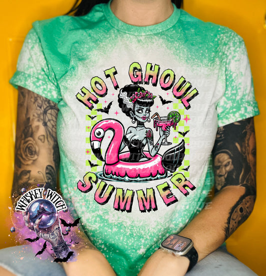 A green and bleach splattered effect  t-shirt with a graphic of a zombie woman in a flamingo pool float, holding a cocktail. The text "Hot Ghoul Summer" is printed on the shirt.