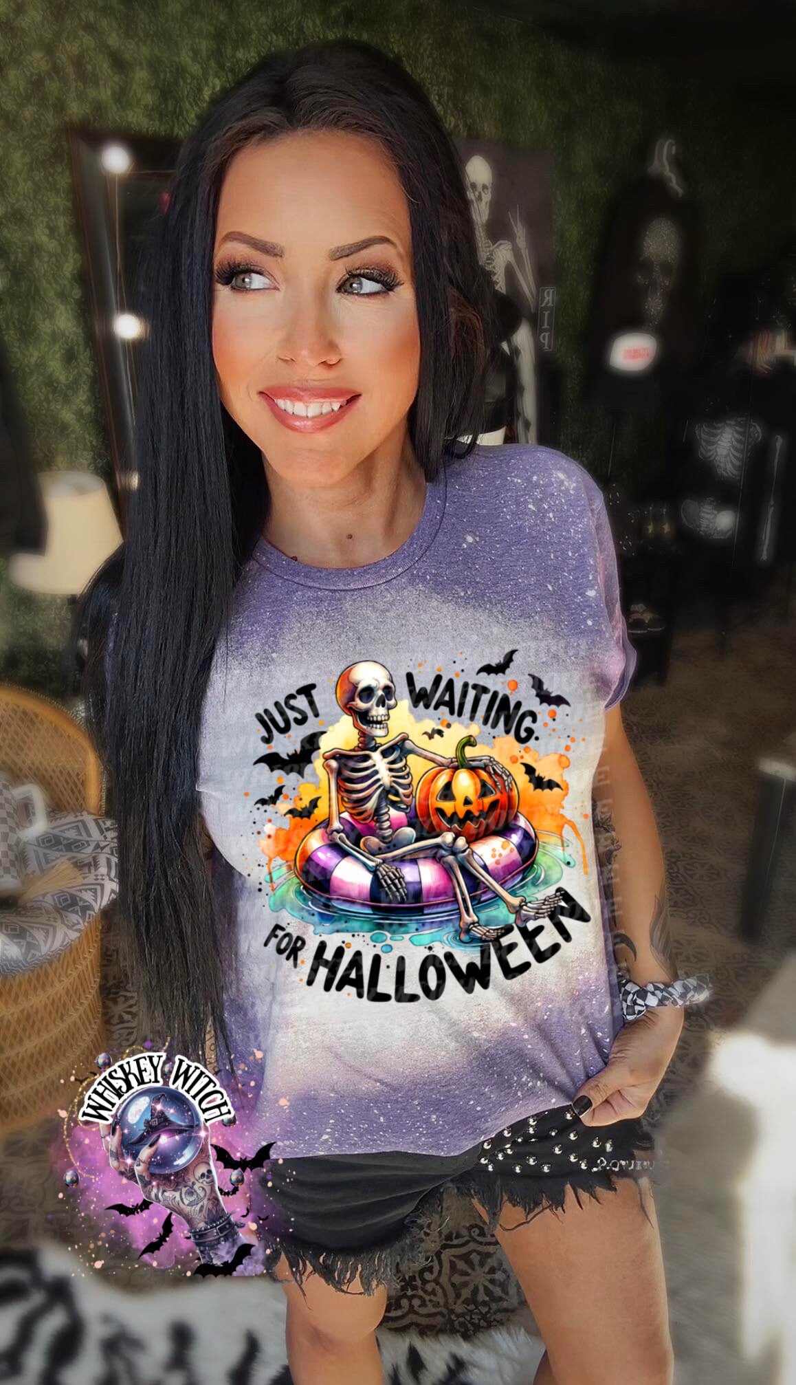 Purple t-shirt with a bleached effect featuring a skeleton sitting in a pool float with a pumpkin. The text on the shirt reads "Just Waiting for Halloween".