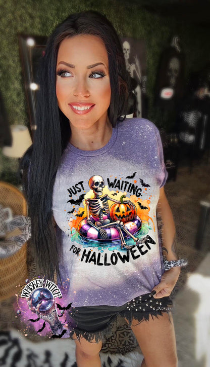 Purple t-shirt with a bleached effect featuring a skeleton sitting in a pool float with a pumpkin. The text on the shirt reads "Just Waiting for Halloween".