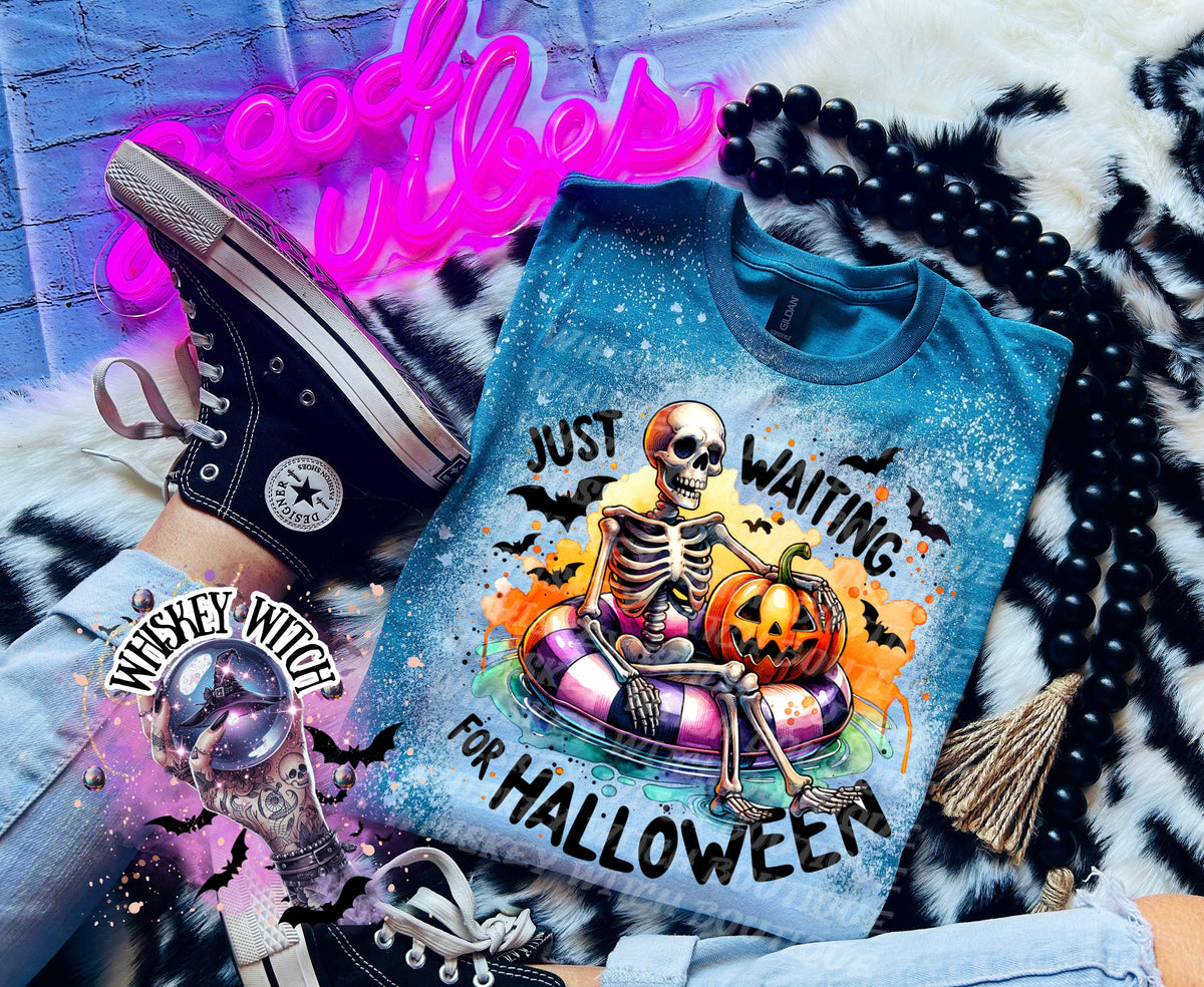 A teal blue t-shirt with a bleach-splattered design featuring a skeleton sitting in a purple and white inner tube with a jack-o-lantern. The text "Just Waiting for Halloween" is printed on the shirt.