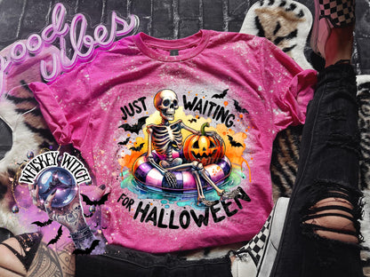 Pink t-shirt with a skeleton wearing a floatie and holding a pumpkin. The text on the shirt reads "Just Waiting for Halloween".