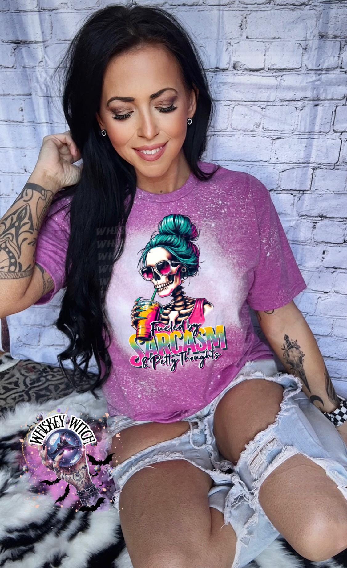 A Berry , bleach tie-dye t-shirt with a graphic of a skeleton wearing sunglasses and a bun, holding a drink. The text on the shirt reads "Fueled by Sarcasm & Petty Thoughts".