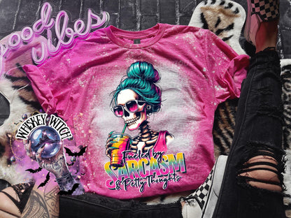 Pink bleached t-shirt with a graphic of a skeleton with blue hair and sunglasses drinking a beverage. The text "Fueled by Sarcasm & Petty Thoughts" is printed on the shirt.