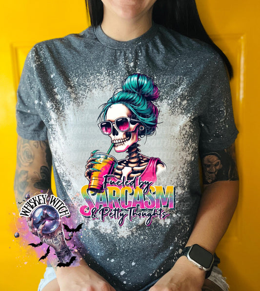 A gray t-shirt with a bleach-splattered design featuring a skeleton with blue hair holding a colorful drink. The text "Fueled by Sarcasm & Petty Thoughts" is printed on the shirt.