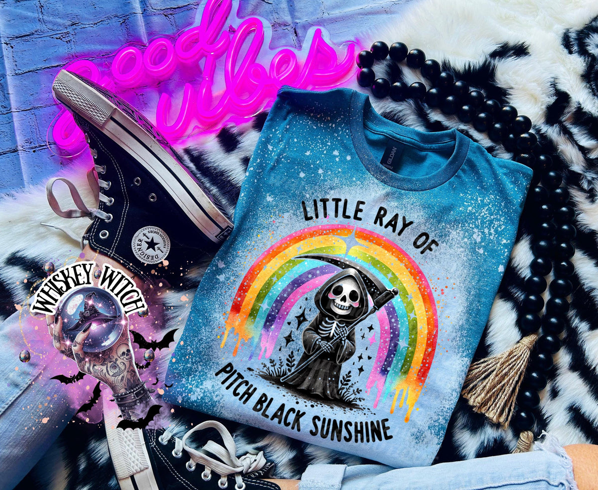 A blue t-shirt with a graphic of a skeleton grim reaper holding a scythe in front of a rainbow. The text "Little Ray of Pitch Black Sunshine" is above and below the graphic. Black Converse sneakers are in the image.