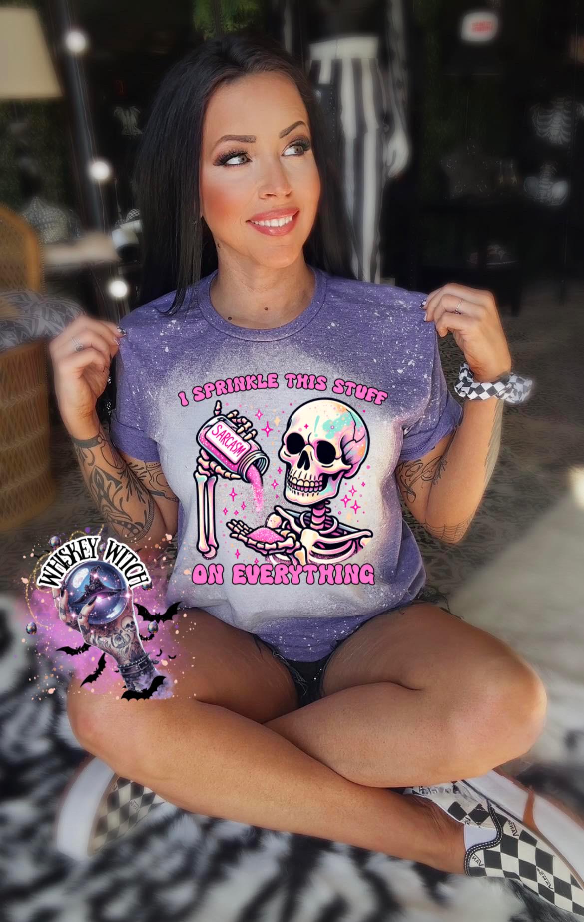 A purple T shirt with bleach splattered design with a colorful skeleton holding a pouring a salt shaker that has the work ‘sarcasm’ on it with the words on the design ‘ I sprinkle this stuff on everything’