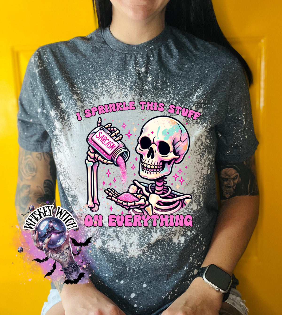 A grey T shirt with bleach splattered design with a colorful skeleton holding a pouring a salt shaker that has the work ‘sarcasm’ on it with the words on the design ‘ I sprinkle this stuff on everything’