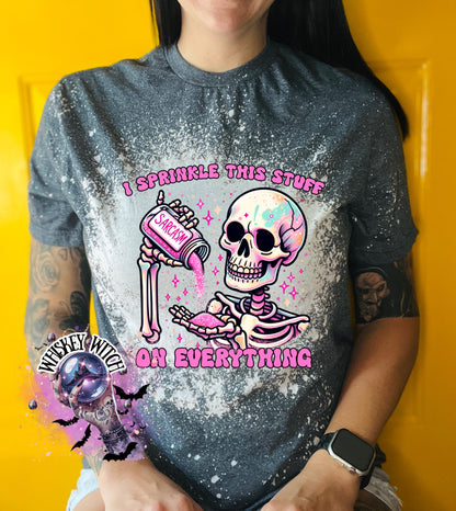 A grey T shirt with bleach splattered design with a colorful skeleton holding a pouring a salt shaker that has the work ‘sarcasm’ on it with the words on the design ‘ I sprinkle this stuff on everything’