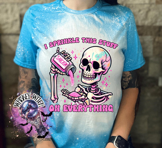 A Saphire blue T shirt with bleach splattered design with a colorful skeleton holding a pouring a salt shaker that has the work ‘sarcasm’ on it with the words on the design ‘ I sprinkle this stuff on everything’