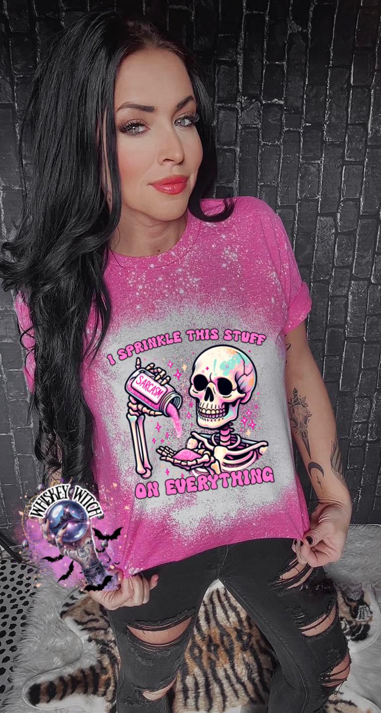 A pink  T shirt with bleach splattered design with a colorful skeleton holding a pouring a salt shaker that has the work ‘sarcasm’ on it with the words on the design ‘ I sprinkle this stuff on everything’