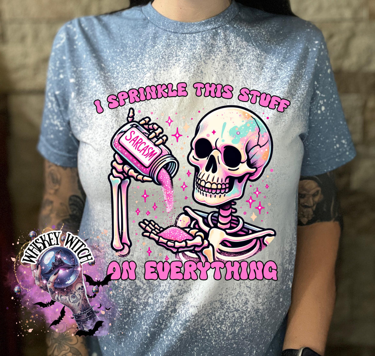 A indigo  blue T shirt with bleach splattered design with a colorful skeleton holding a pouring a salt shaker that has the work ‘sarcasm’ on it with the words on the design ‘ I sprinkle this stuff on everything’