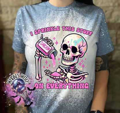 A indigo  blue T shirt with bleach splattered design with a colorful skeleton holding a pouring a salt shaker that has the work ‘sarcasm’ on it with the words on the design ‘ I sprinkle this stuff on everything’