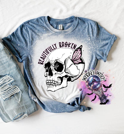 Indigo blue colored  t-shirt with a white bleached design featuring a skull with a pink butterfly on its head and the text "Beautifully Broken". The design is surrounded by a pink and purple watercolor effect.