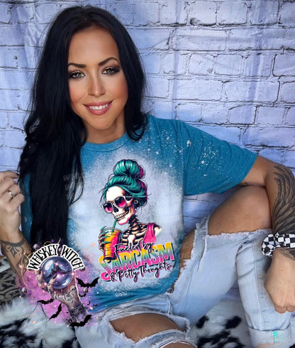 A teal blue t-shirt with a bleached effect featuring a skeleton with a messy bun, sunglasses, and a pink tank top holding a drink. The text on the shirt reads "Fueled by Sarcasm & Petty Thoughts".