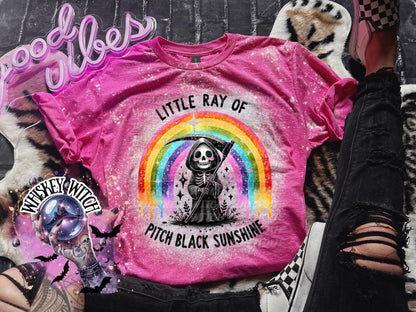 : Pink t-shirt with a distressed design featuring a rainbow and a Grim Reaper character holding a scythe. The text reads "LITTLE RAY OF PITCH BLACK SUNSHINE".