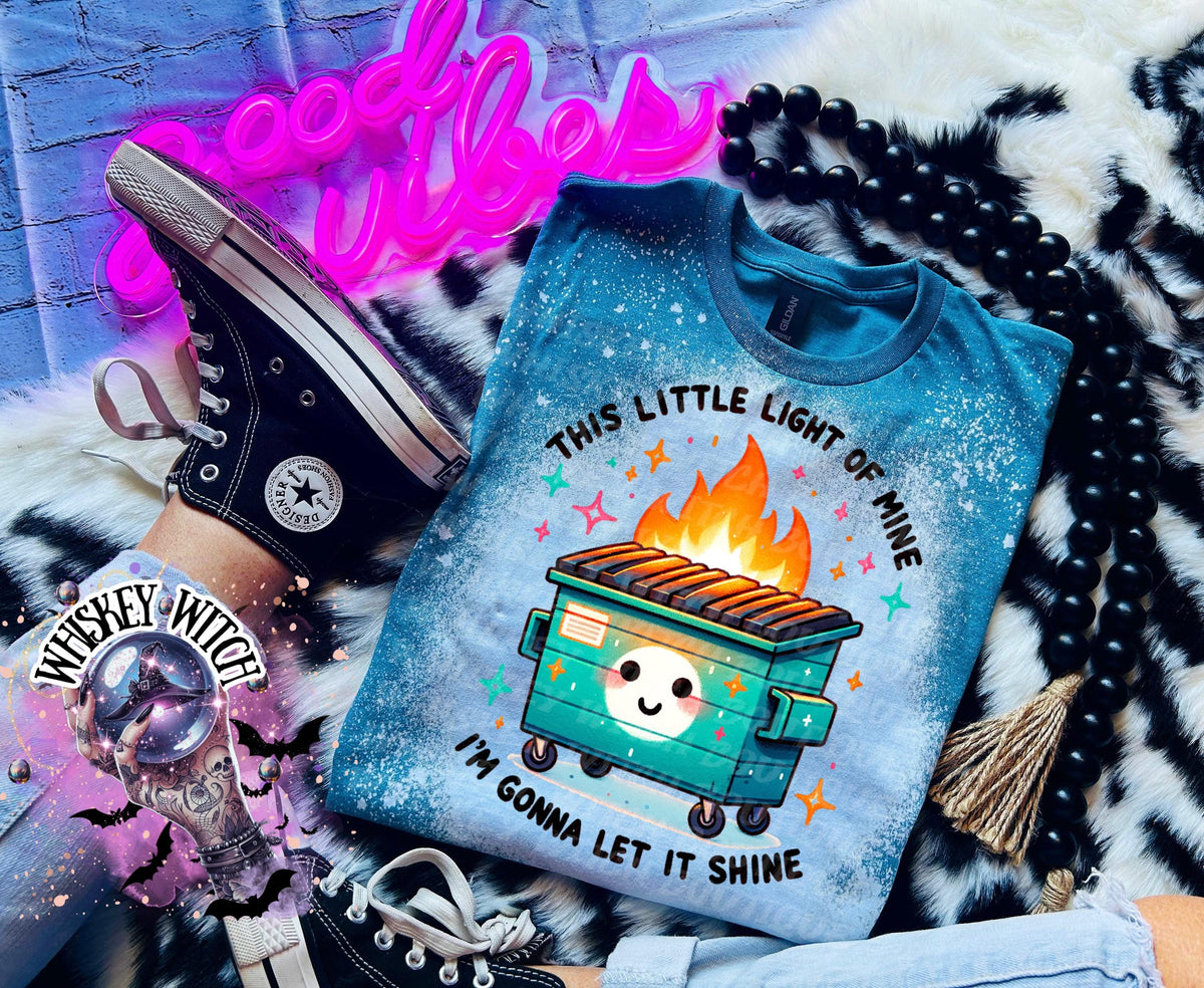 A teal blue t-shirt with a bleached effect featuring a cartoon image of a green dumpster on fire with the text "This little light of mine, I'm gonna let it shine".