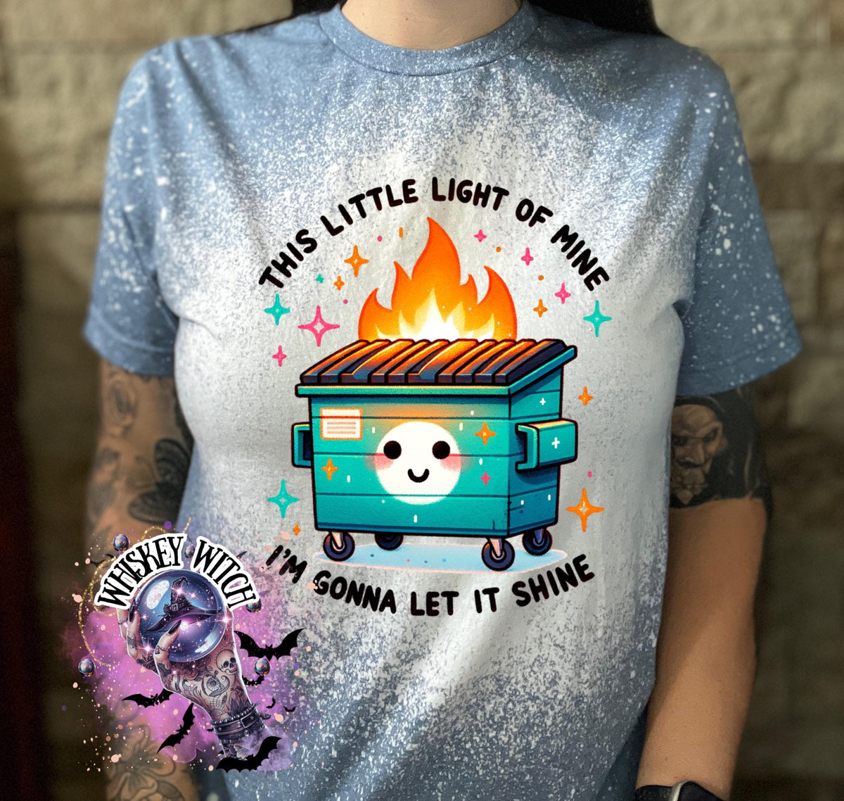 A light blue t-shirt with a bleach-splattered design. The shirt features a cartoon image of a smiling trash can with flames coming out of the top. The text "This little light of mine I'm gonna let it shine" is printed on the shirt.