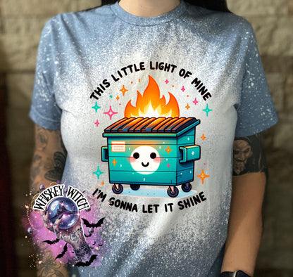 A light blue t-shirt with a bleach-splattered design. The shirt features a cartoon image of a smiling trash can with flames coming out of the top. The text "This little light of mine I'm gonna let it shine" is printed on the shirt.