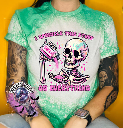A green T shirt with bleach splattered design with a colorful skeleton holding a pouring a salt shaker that has the work ‘sarcasm’ on it with the words on the design ‘ I sprinkle this stuff on everything’
