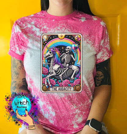 A pink  t-shirt with a bleached design featuring a tarot card with the image of  skeleton holding a glass of wine  riding a unicorn with a rainbow behind them with the words of “The Audacity” at the bottom