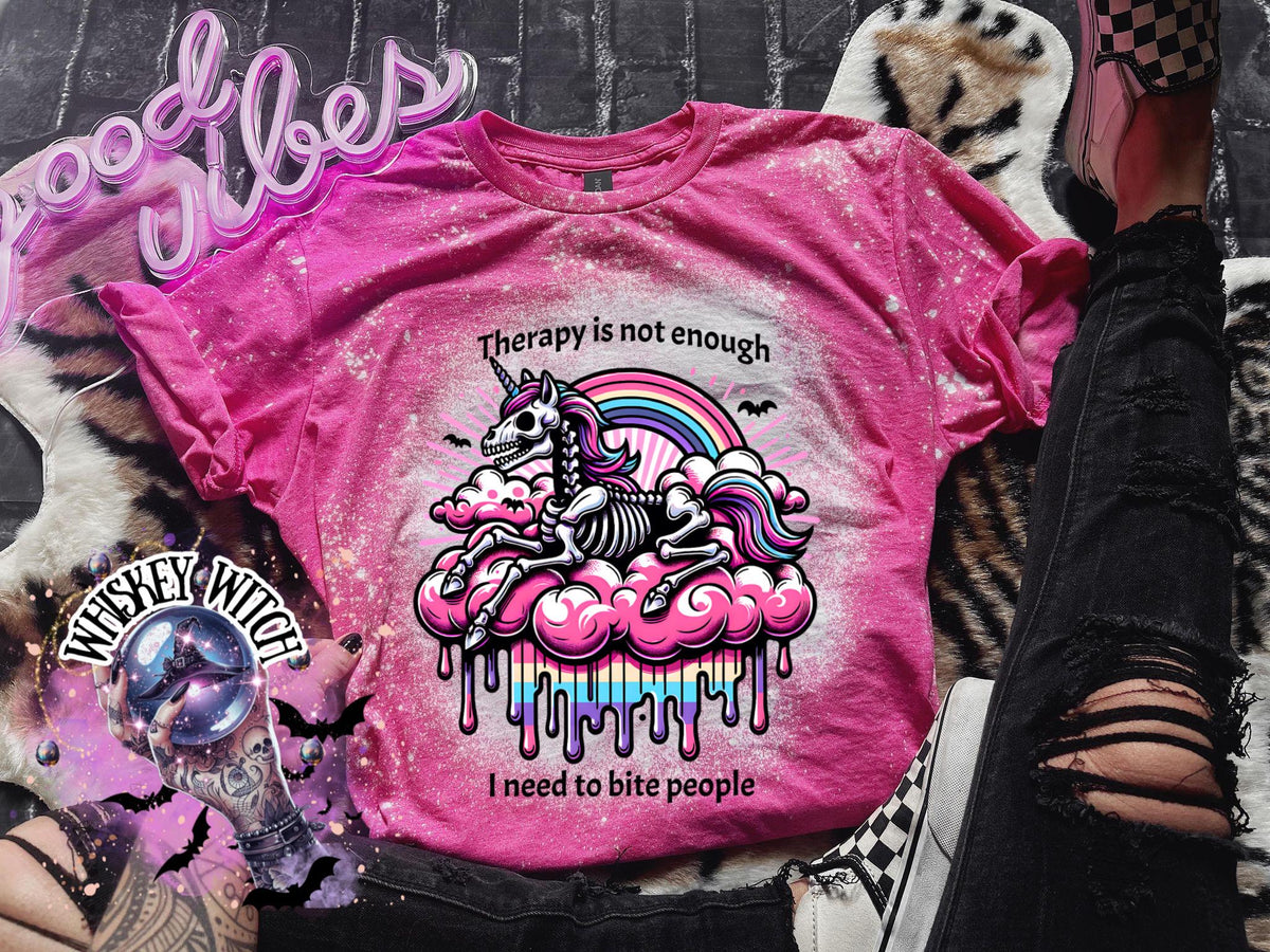 A pink and bleached design T shirt with a skeleton unicorn and a rainbow with a dripping colored cloud with the words “Therapy is not enough, I need to bite people.”