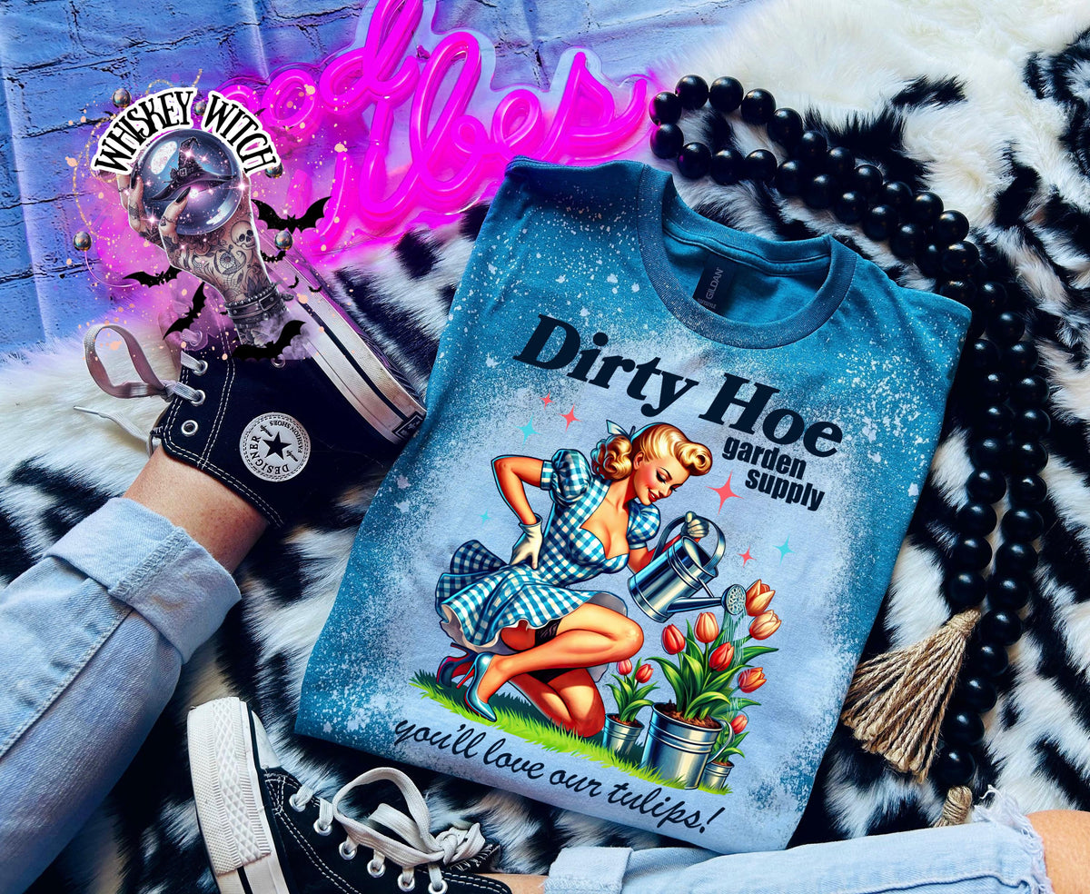 A blue t-shirt with a bleach  white splatter design featuring a woman in a gingham dress holding a watering can. The text on the shirt reads "Dirty Hoe garden supply you'll love our tulips!"