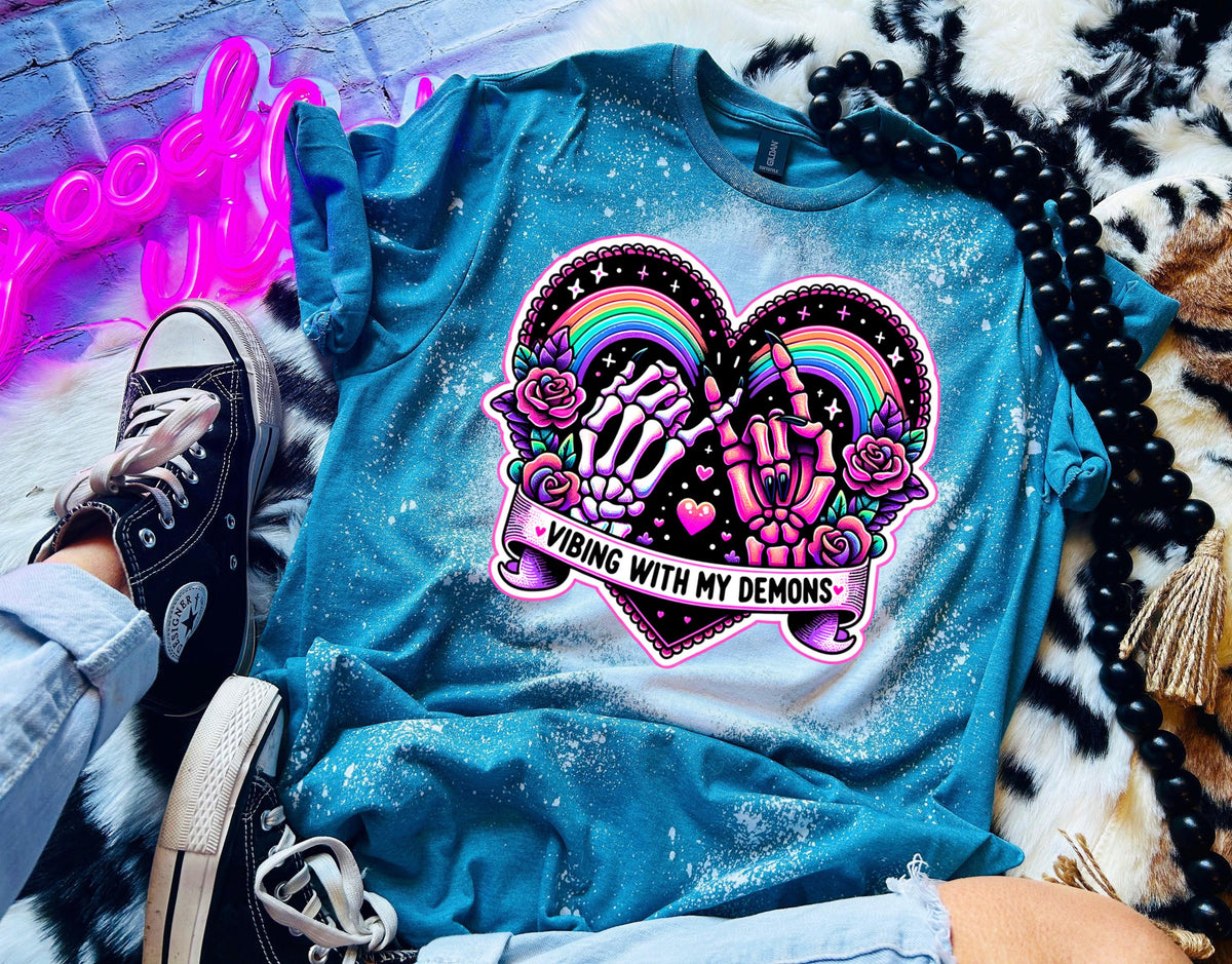 A teal blue t-shirt with a white bleach design of a heart with a rainbow, two skeleton hands, and flowers. The text 'Vibing With My Demons' is written below the heart.