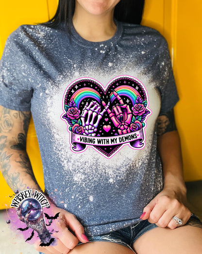 A gray t-shirt with a bleach-splattered design featuring a pink heart with a rainbow and two hands, one skeleton and one with claws, holding pinky fingers together. The text 'Vibing with my demons' is written below the heart.