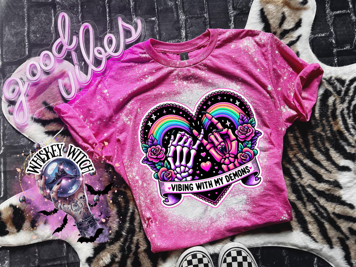 Pink bleached t-shirt with a black heart graphic. The heart graphic features two skeleton hands holding a rainbow with the text 'Vibing with my demons'.