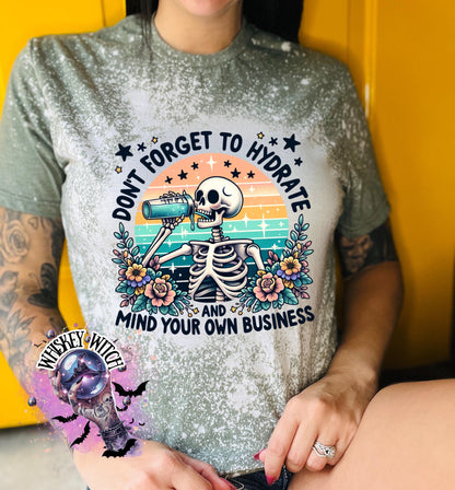 A military  green t-shirt with a bleached effect features a skeleton drinking from a blue bottle with the text "Don't Forget to Hydrate and Mind Your Own Business" in a retro sunset design.