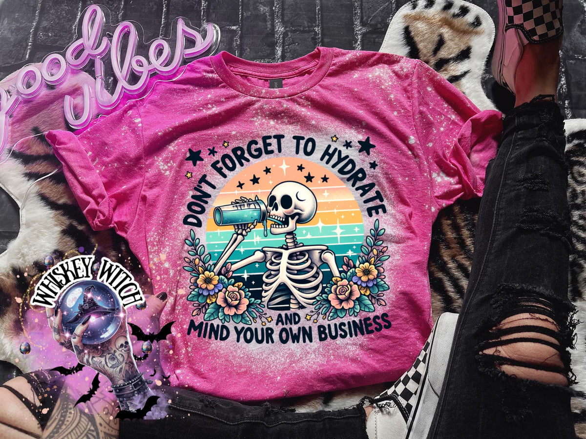 Pink t-shirt with a bleached effect featuring a skeleton drinking from a blue bottle with the text "Don't forget to hydrate and mind your own business".