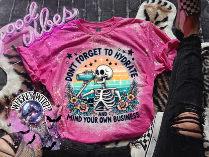 Pink t-shirt with a bleached effect featuring a skeleton drinking from a blue bottle with the text "Don't forget to hydrate and mind your own business".