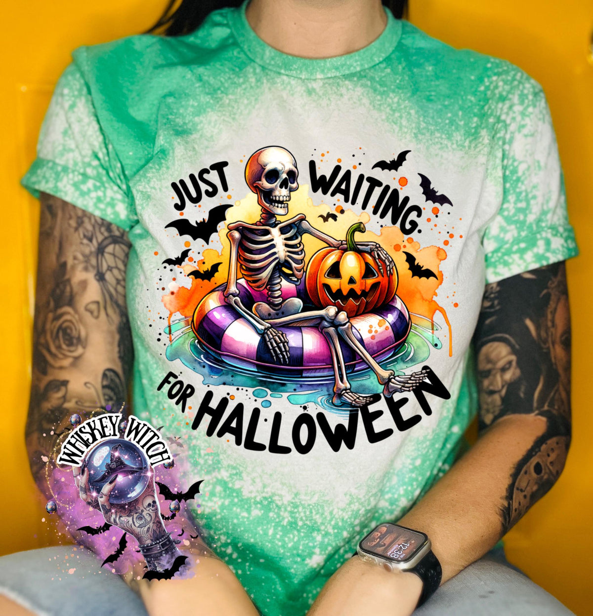Green t-shirt with a bleached effect featuring a skeleton sitting in a pool float with a pumpkin. The text on the shirt reads "Just Waiting for Halloween".