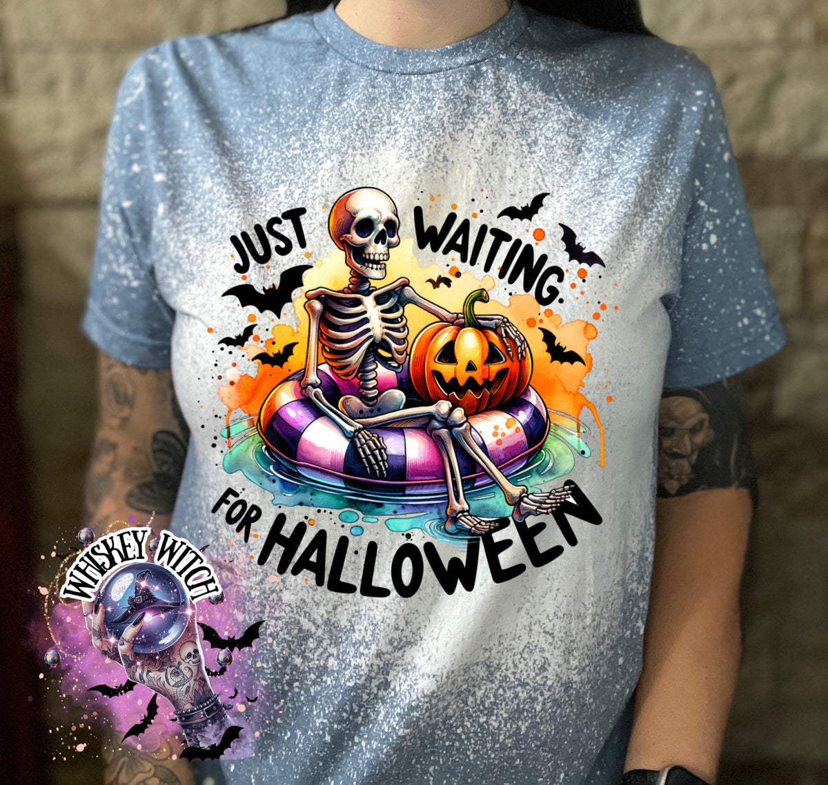 Indigo Blue  t-shirt with a bleached effect featuring a skeleton sitting in a pool float with a pumpkin. The text on the shirt reads "Just Waiting for Halloween".