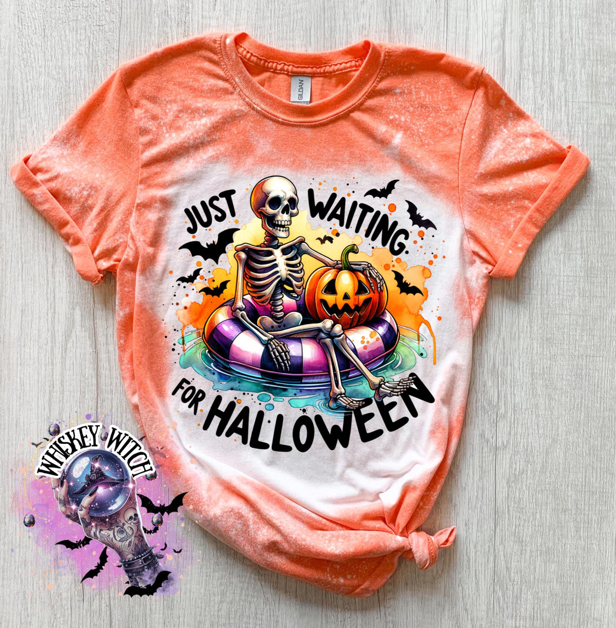 Orange t-shirt with a bleached effect featuring a skeleton sitting in a pool float with a pumpkin. The text on the shirt reads "Just Waiting for Halloween".