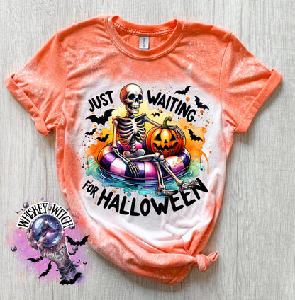Orange t-shirt with a bleached effect featuring a skeleton sitting in a pool float with a pumpkin. The text on the shirt reads "Just Waiting for Halloween".