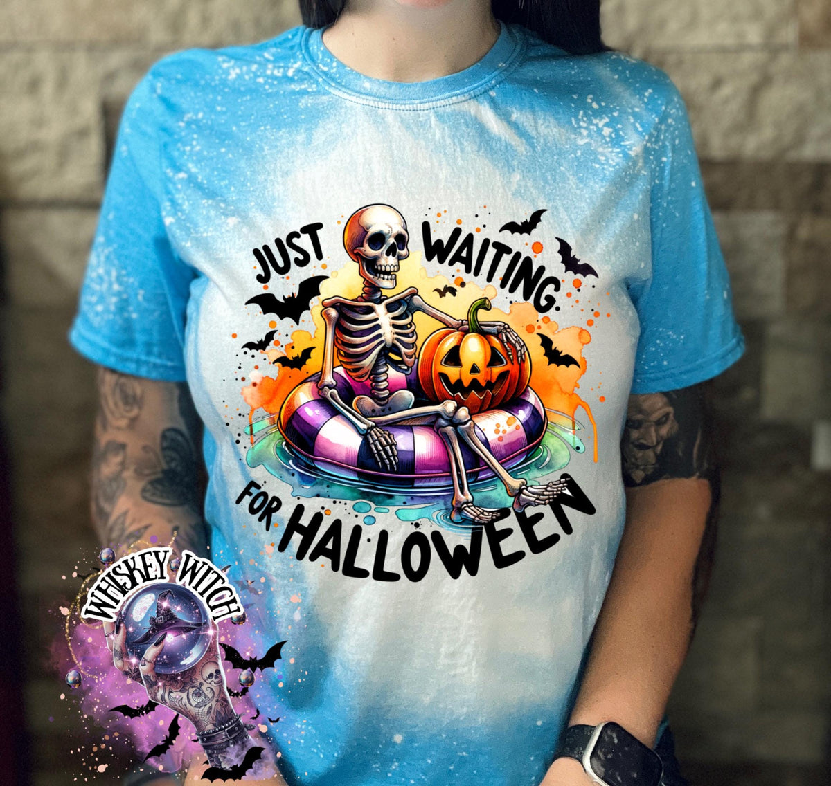 Saphire Blue t-shirt with a bleached effect featuring a skeleton sitting in a pool float with a pumpkin. The text on the shirt reads "Just Waiting for Halloween".