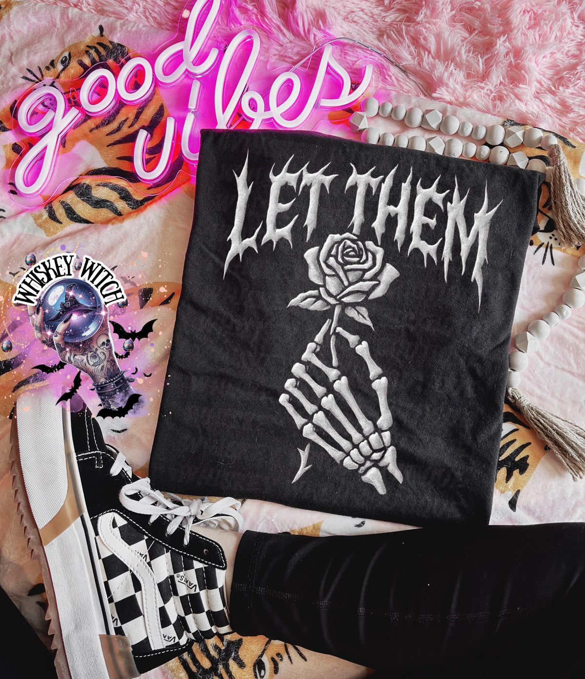 Black t-shirt with the text "LET THEM" in white gothic lettering. A white skeleton hand holds a white rose. The image also includes a pair of black and white checkered sneakers, a neon sign that says "good vibes", and a decorative light.