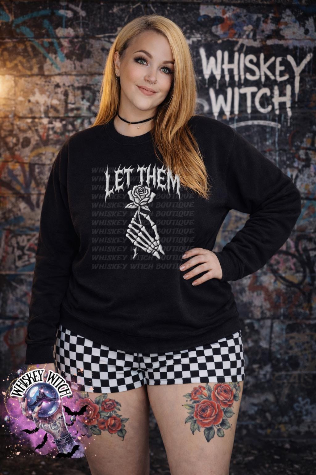 Black sweatshirt with the text "LET THEM" and a skeleton hand holding a rose. The words "Whiskey Witch Boutique" are printed on the shirt. The model is wearing black and white checkered shorts.