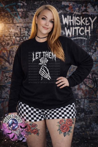Black sweatshirt with the text "LET THEM" and a skeleton hand holding a rose. The words "Whiskey Witch Boutique" are printed on the shirt. The model is wearing black and white checkered shorts.