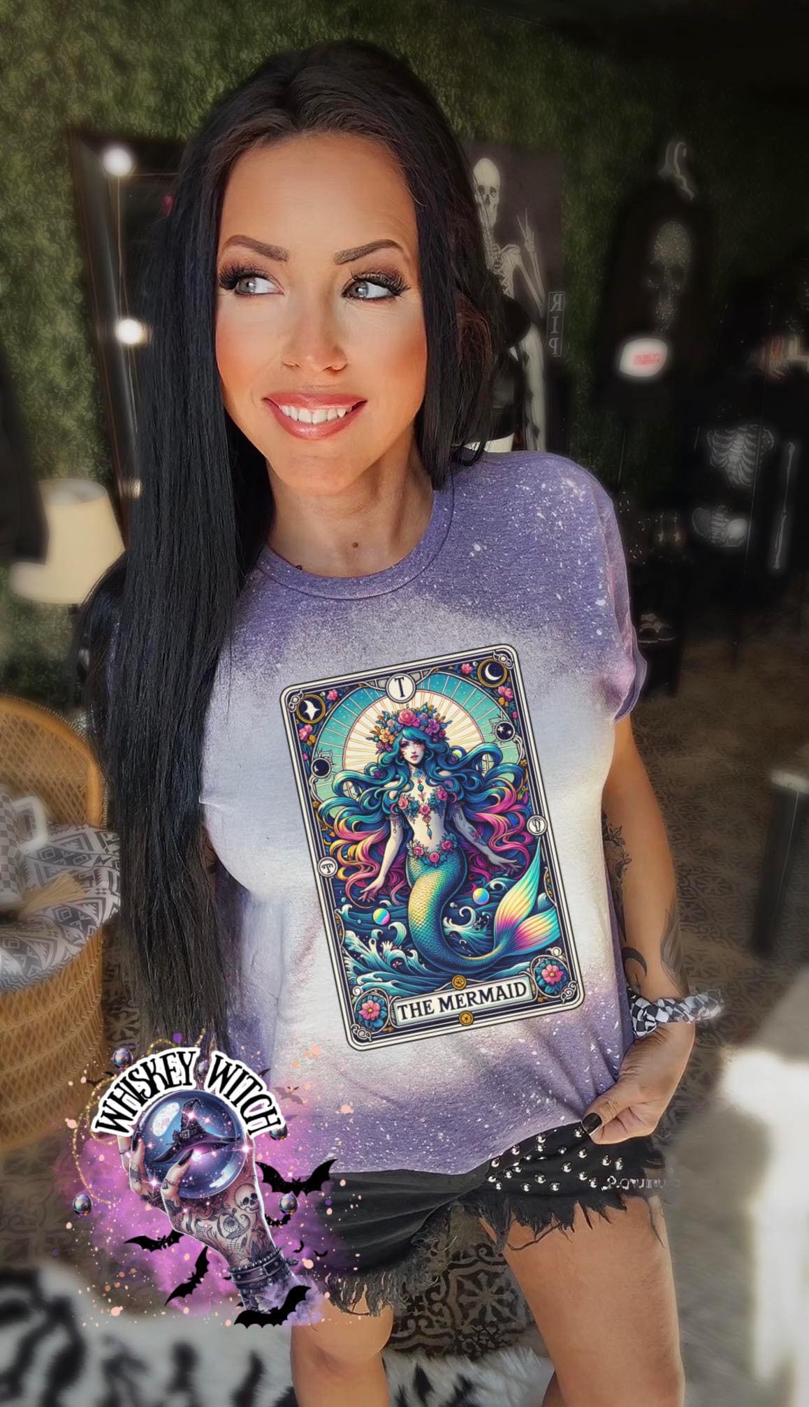 Mystical mermaid tarot card graphic centered on a purple bleach-dyed shirt, photographed with neon pink sign and cozy background props.