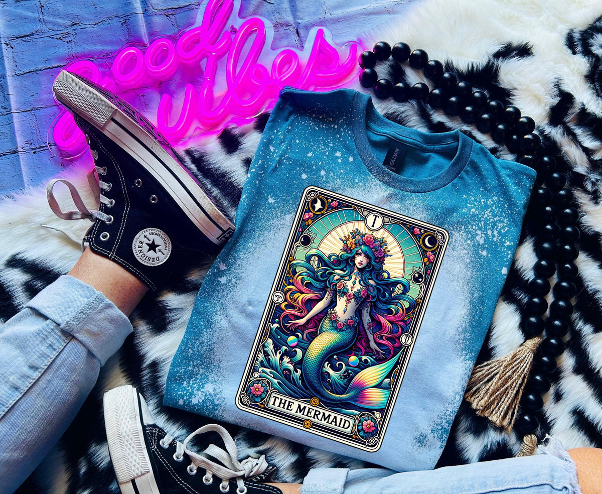 Mystical mermaid tarot card graphic centered on a Galapagos blue bleach-dyed shirt, photographed with neon pink sign and cozy background props.
