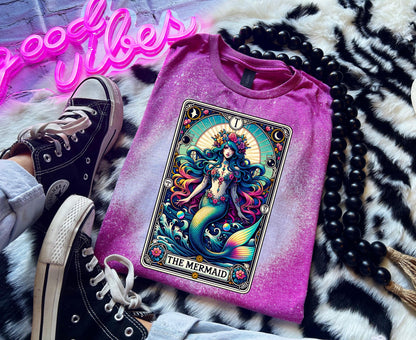 Mystical mermaid tarot card graphic centered on a Berry pink  bleach-dyed shirt, photographed with neon pink sign and cozy background props.