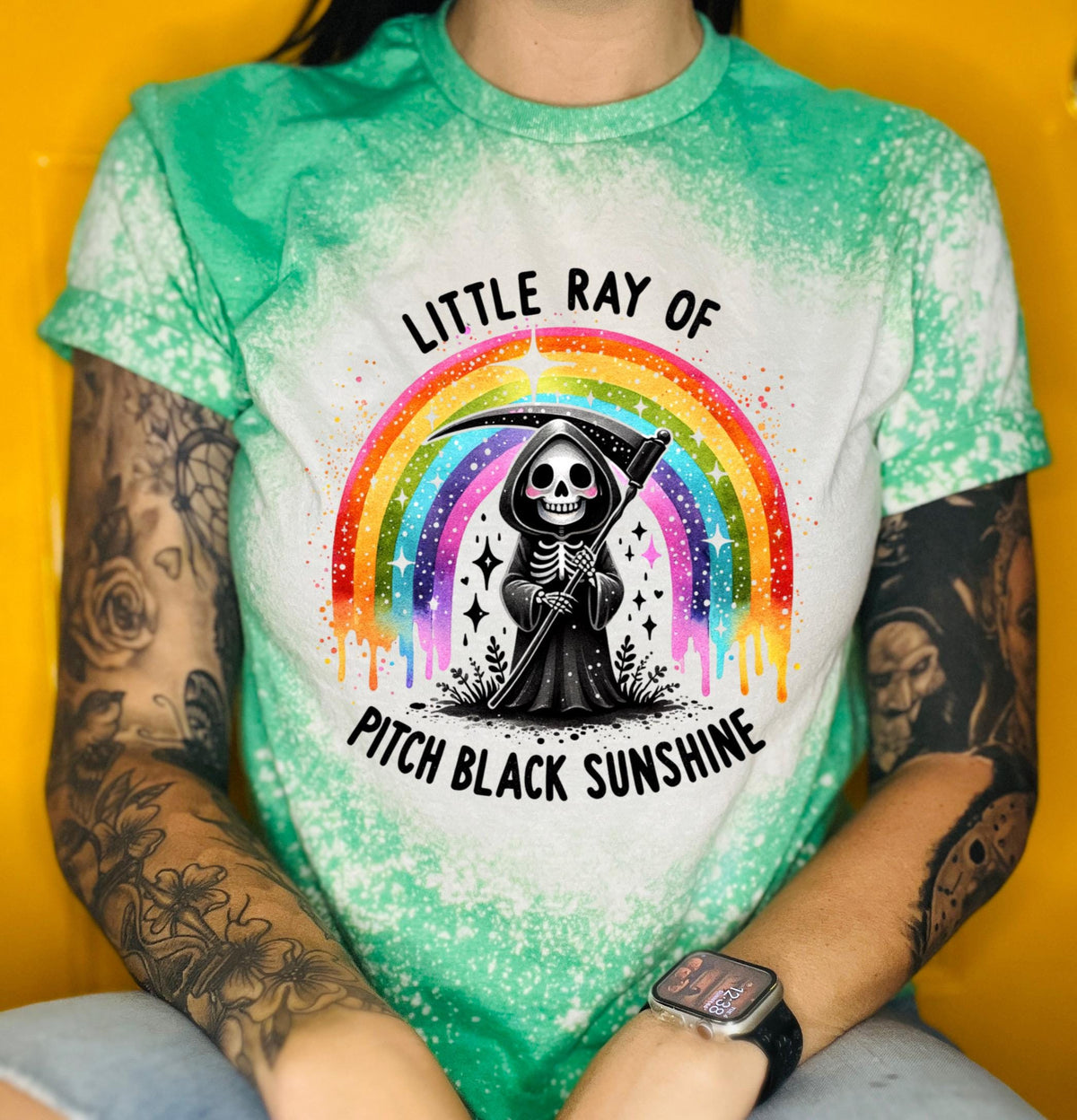 Close-up of bleach-wash tee showing dripping rainbow over a skeleton reaper holding a scythe with “Little Ray Of Pitch Black Sunshine” text.