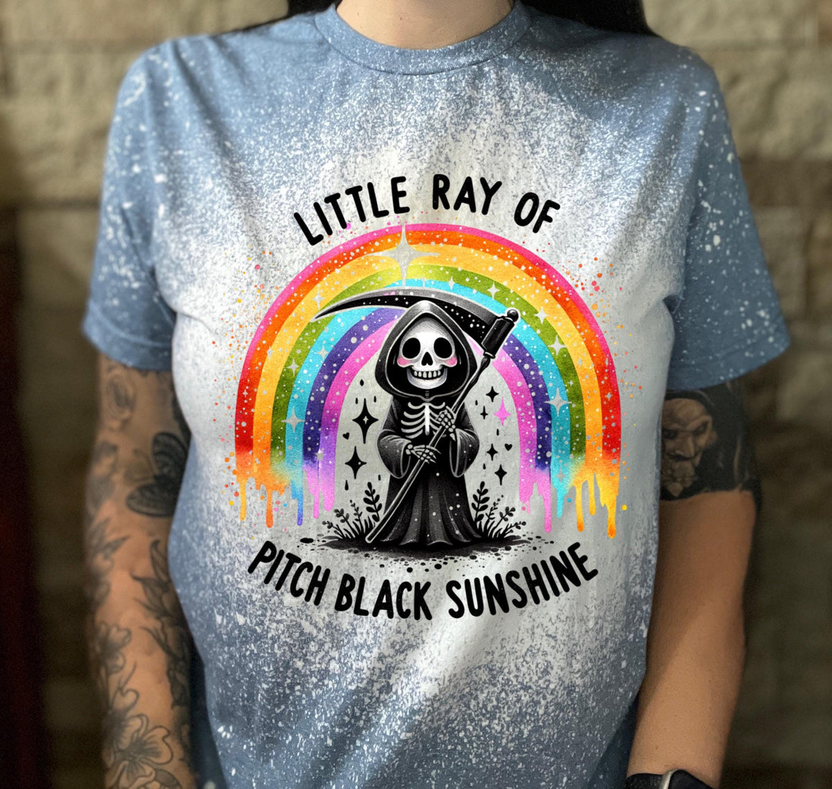 Close-up of bleach-wash tee showing dripping rainbow over a skeleton reaper holding a scythe with “Little Ray Of Pitch Black Sunshine” text.